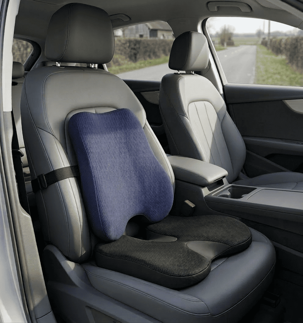 Photo de l'intérieur d'une voiture moderne montrant un kit de confort ergonomique installé sur le siège conducteur en cuir gris. L'ensemble comprend un coussin lombaire bleu marine avec sangle de fixation noire sur le dossier et un coussin d'assise noir à mémoire de forme avec découpe en U pour le coccyx. La vue est prise depuis la portière ouverte, avec une route visible en arrière-plan à travers le pare-brise.